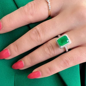 May include: A silver ring with a large emerald green gemstone surrounded by smaller diamonds. The ring is on a finger with pink nail polish.