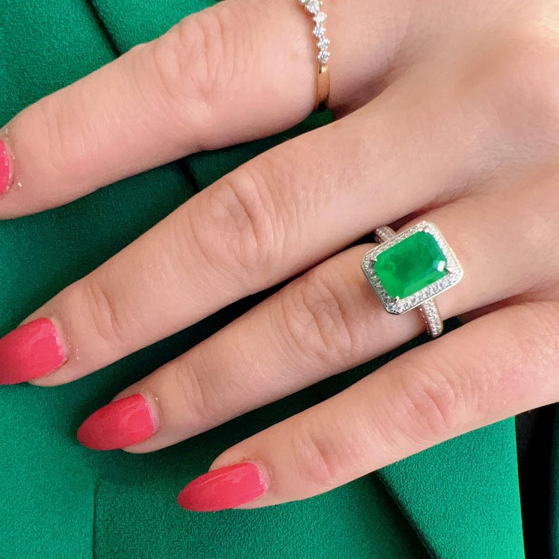 May include: A silver ring with a large emerald green gemstone surrounded by smaller diamonds. The ring is on a finger with pink nail polish.