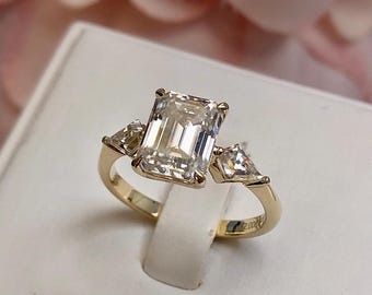 May include: A gold engagement ring with a large emerald cut diamond and two smaller triangular diamonds on the sides.