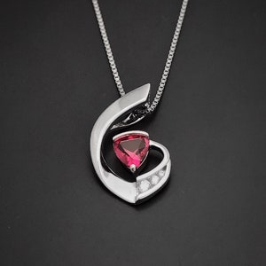 May include: A silver pendant necklace featuring a red gemstone at its centre, surrounded by smaller clear gemstones. The pendant is shaped like a swirl.
