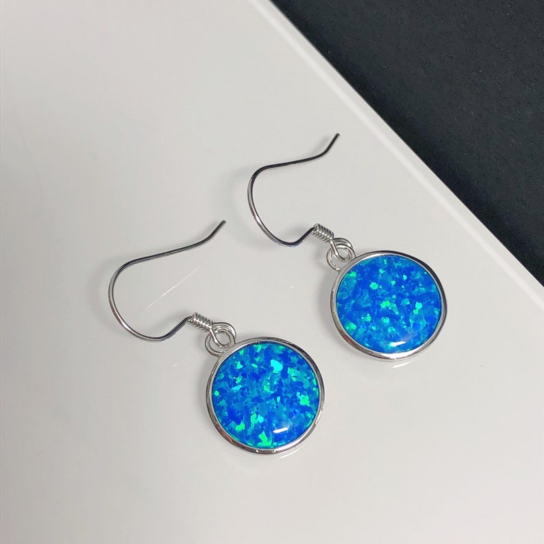 May include: A pair of silver-toned dangle earrings. Each earring features a round, bezel-set, vibrant blue opal stone with flecks of green. The earrings have a simple hook design for pierced ears.
