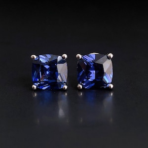 May include: A pair of square-cut, deep blue gemstone stud earrings. The gems are set in a silver-colored metal with four prongs. The earrings are displayed against a dark background, highlighting their color and sparkle.