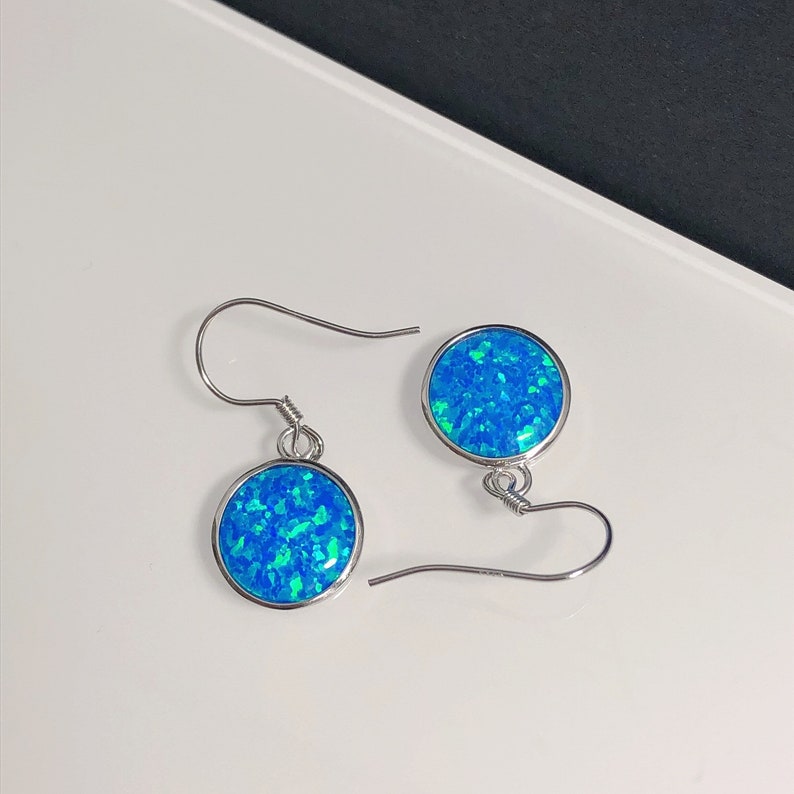 May include: A pair of silver-toned dangle earrings. Each earring features a round, bezel-set, blue opal stone. The opal has a mosaic appearance with shades of blue and green. The earrings have a simple hook closure.