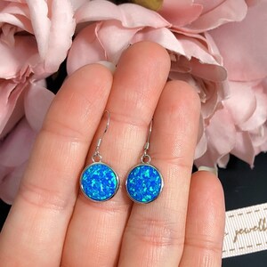 May include: A pair of round, blue opal earrings with a silver-colored border. The earrings are held in a hand, with a soft pink floral background. The opal has a speckled appearance, with shades of blue and turquoise.