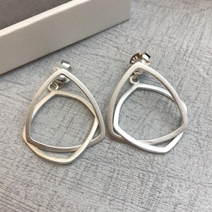 May include: A pair of silver earrings. Each earring has a geometric design with a triangular top and a square shape below. The earrings are shown on a grey surface, with a white box in the background.