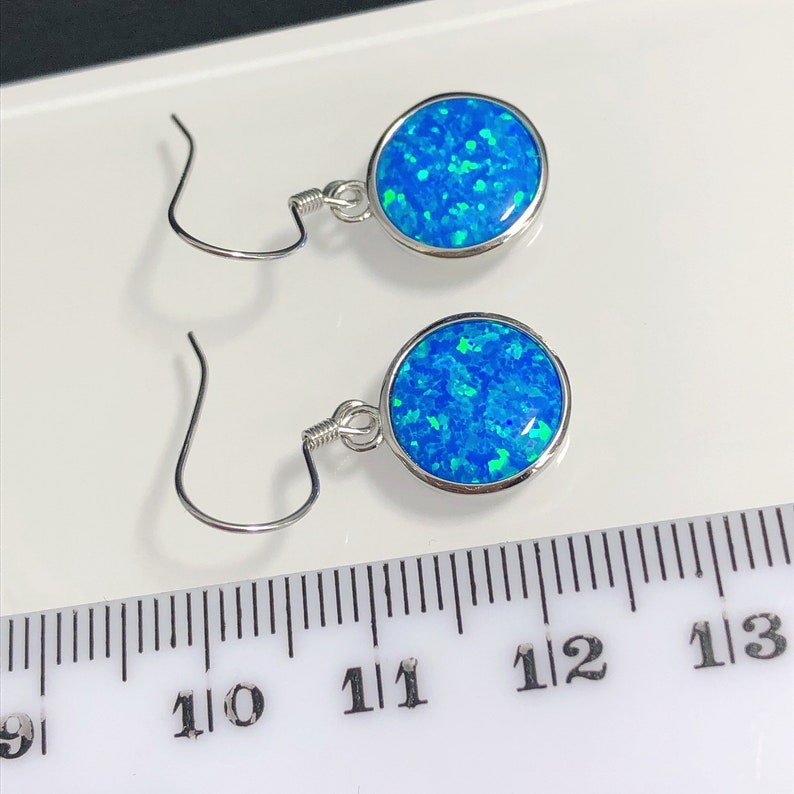 May include: A pair of silver-toned earrings with round, blue opal stones. The opals have a speckled appearance with shades of blue and green. The earrings are displayed on a white surface next to a ruler.