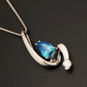 May include: A silver pendant necklace with a teardrop-shaped blue gemstone and a small, round, clear gemstone. The pendant has a curved, abstract design. The necklace chain is a fine, silver box chain. The background is black.