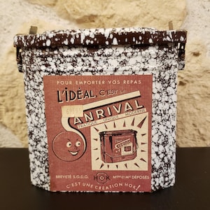 Op de afbeelding: Een witte en bruine gevlekte metalen lunchbox met een rood en wit label waarop staat "L'IDEAL, C'EST LA ANRIVAL PRATIQUE UNIQUE MODERNE" en "BREVETÉ S.G.D.G. HOK MARQUE ET MODELE DEPOSES C'EST UNE CREATION HOK!"