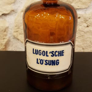 Puede incluir: Una botella de vidrio ámbar vintage con un tapón oscuro. La botella tiene una etiqueta blanca con el texto azul "LUGOL'SCHE L'OSUNG". La botella es probablemente para almacenar líquidos. El fondo es una pared de piedra.