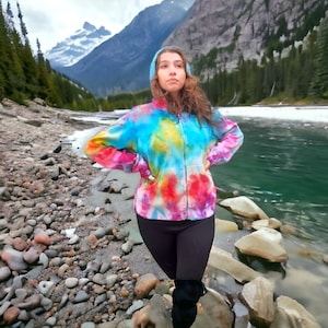 May include: A person wearing a colorful tie-dye hoodie with a zipper and black pants, standing in front of a river and mountains.