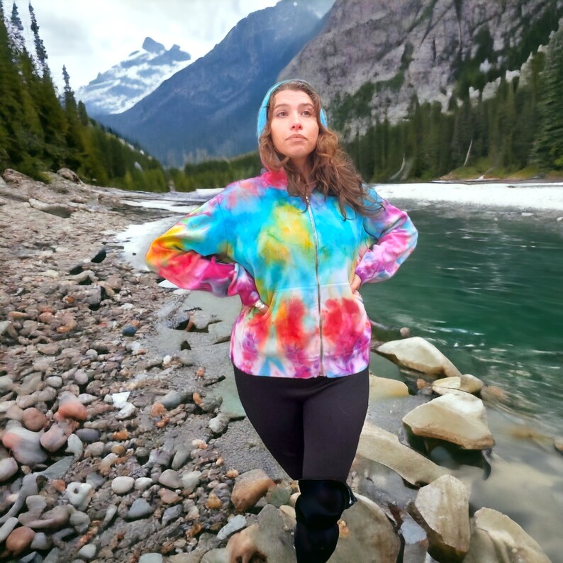 May include: A person wearing a colorful tie-dye hoodie with a zipper and black pants, standing in front of a river and mountains.
