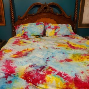 May include: A bedding set featuring a tie-dye pattern in vibrant shades of bright pink, sunny yellow, and sky blue. The set includes a large comforter and two matching pillowcases, all displaying a swirling, multi-colored abstract design.