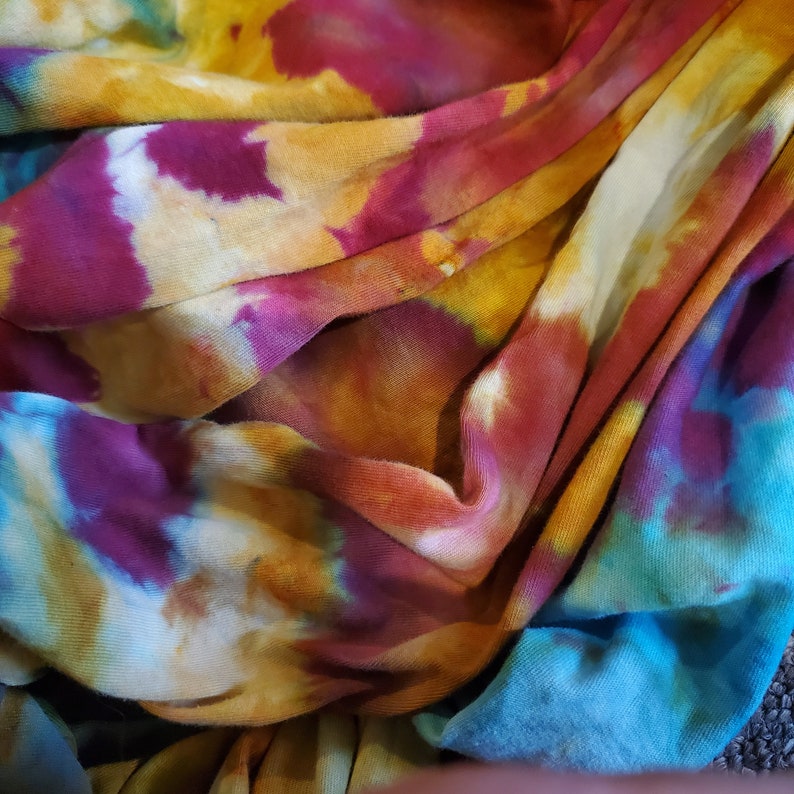 K&ouml;nnte beinhalten: Nahaufnahme eines Batik-Stoffes in Blaut&ouml;nen, Lila, Gelb und Orange. Der Stoff ist gefaltet und drapiert und erzeugt ein strukturiertes und farbenfrohes Muster.