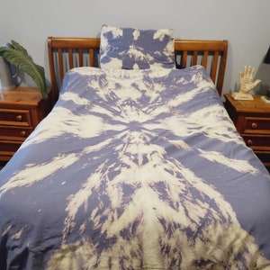 May include: A blue and white tie-dye duvet cover with a circular pattern. The duvet cover is on a wooden bed frame with a matching tie-dye pillow.