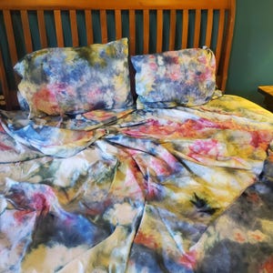 May include: A set of bed linens featuring a tie-dye pattern with blended colors of deep blue, soft pink, muted yellow, and charcoal gray. The set includes two rectangular pillowcases and a matching flat sheet, all displaying a swirling, abstract design.