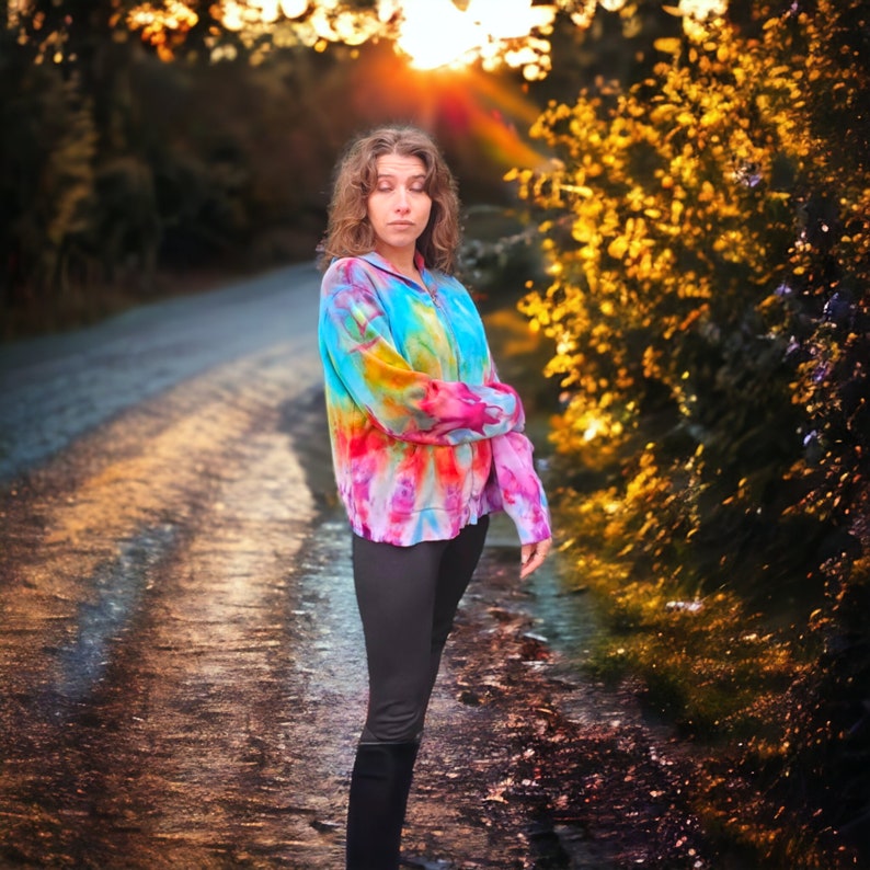May include: A woman wearing a colorful tie-dye sweatshirt and black pants stands on a paved road. The road is lined with trees and bushes. The sun is shining brightly in the background.