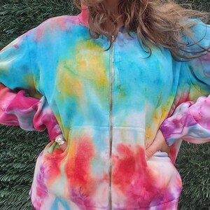 May include: A tie-dye zip-up hoodie with a blue, pink, yellow, and white color scheme. The hoodie has a front pocket and a zipper closure.