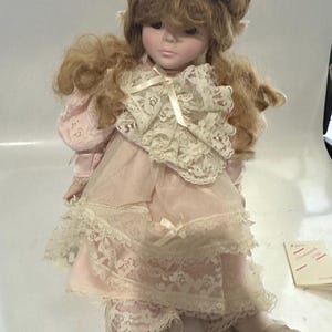 Może przedstawiać: Zabytkowa porcelanowa lalka z blond włosami ułożonymi w kok i kaskadowymi lokami. Lalka ma na sobie różową sukienkę z koronkowym wykończeniem i pasującym koronkowym kołnierzem. Lalka siedzi z wyciągniętymi nogami.