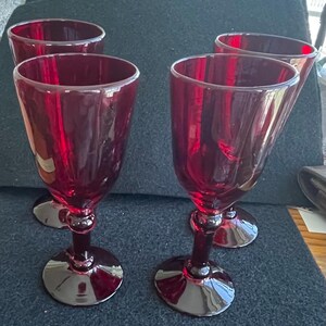 May include: Four ruby red wine glasses. Each glass has a rounded bowl, a bulbous stem, and a circular base. The glasses are made of a transparent material, allowing light to pass through, and are arranged on a dark surface.