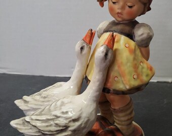 May include: A ceramic figurine of a young girl with closed eyes, wearing a red cap and yellow dress, standing with two white geese. The geese have orange beaks and are positioned on a cream-colored base.