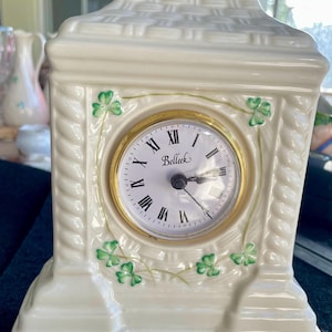 May include: A white porcelain clock with a gold trim and green shamrock accents. The clock face has Roman numerals and says "Belleek" in gold lettering.