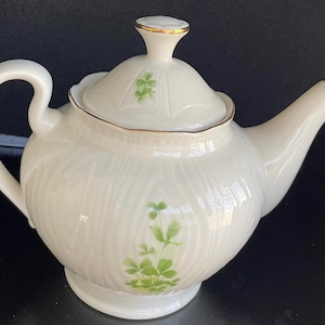 May include: A white porcelain teapot with a gold trim and green shamrock designs. The teapot has a lid with a knob on top.