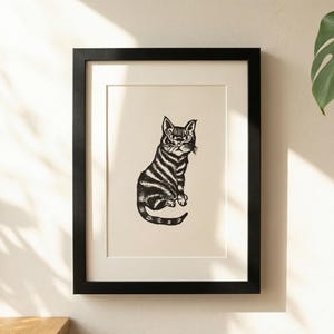 Puede incluir: Una impresión en blanco y negro enmarcada de un gato sentado con marcas rayadas. La obra de arte se muestra en un marco negro con un passe-partout blanco, montada en una pared de color claro. La ilustración del gato tiene patrones de pelaje detallados.