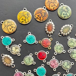 Op de afbeelding: Een verzameling kleine metalen bedels met in reliëf gemaakte cherubijnfiguren in diverse kleuren zoals rood, geel, groen, roze en turkoois. De bedels zijn rond of ovaal van vorm en hebben enkele of dubbele ringetjes voor bevestiging.