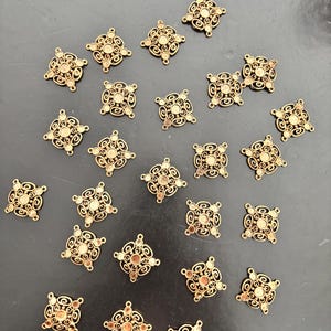 Op de afbeelding: Een verzameling van tweeëntwintig vierkante metalen bedels in een bronzen tint. Elke bedel heeft een ingewikkeld opengewerkt filigraanontwerp met een centrale ronde zetting en vier kleine lusjes op de hoeken voor bevestigingsdoeleinden.