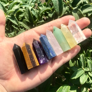 Op de afbeelding: Een set van acht kleine kristallen punten in verschillende kleuren, waaronder zwart, goud, paars, blauw, roze, groen, geel en helder. De punten zijn in een rij op een hand gerangschikt.