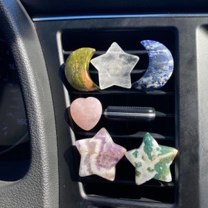 May include: A collection of seven crystal car air freshener charms in the shapes of stars, moons, and hearts. The charms are made of various types of crystals, including amethyst, rose quartz, and green aventurine.