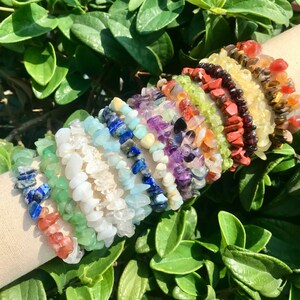 Puede incluir: Un conjunto de 12 pulseras hechas con varias piedras semipreciosas, incluyendo amatista, aguamarina, cornalina, cuarzo transparente, citrino, aventurina verde, lapislázuli, jaspe rojo, ojo de tigre y howlite blanca. Las pulseras están apiladas sobre una superficie blanca.