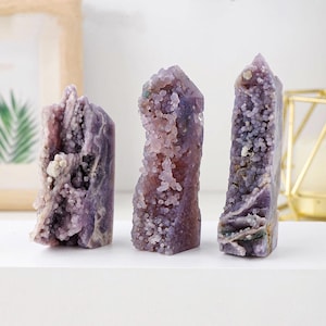 May include: Three amethyst crystal towers in varying shapes and sizes. The crystals display a range of purple hues, from light lavender to deep violet, with a textured, bubbly surface. The crystals are displayed on a white surface.
