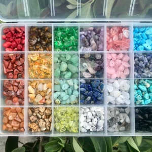 May include: A clear plastic storage box with 16 compartments filled with various types of small, colorful gemstone beads. The beads are arranged in rows and columns, and each compartment has a different color of beads.