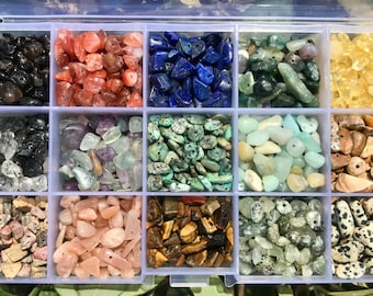 Puede incluir: Una caja de almacenamiento de plástico transparente llena de 12 compartimentos, cada uno con un tipo diferente de pequeña piedra preciosa pulida. Las piedras preciosas son de varios colores, incluyendo negro, rojo, azul, verde, blanco, rosa, marrón y gris.