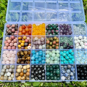 Op de afbeelding: Een transparante plastic opbergdoos gevuld met verschillende ronde kralen in verschillende kleuren. De kralen zijn gerangschikt in rijen en kolommen, waarbij elk vakje een ander type kraal bevat. De kralen zijn gemaakt van verschillende materialen, waaronder glas, steen en hout.