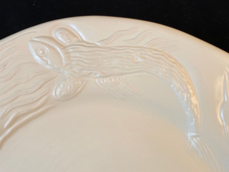 K&ouml;nnte beinhalten: Wei&szlig;e Keramikplatte mit einem erhabenen Design eines Fisches, der im Wasser schwimmt. Der Fisch ist mit Schuppen und Flossen detailliert.