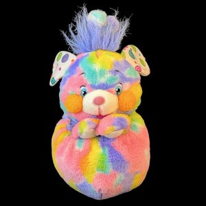 Puede incluir: Un peluche con un patrón tie-dye multicolor en rosa, azul, amarillo y verde. La criatura tiene un mechón de pelo morado en la parte superior, grandes ojos azules, mejillas naranjas y orejas con un patrón de espiral colorido en el interior.