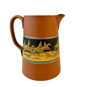 Puede incluir: Una jarra de cerámica marrón con un diseño de escena de caza en oro y negro. La escena presenta jinetes a caballo y perros que persiguen a un zorro.