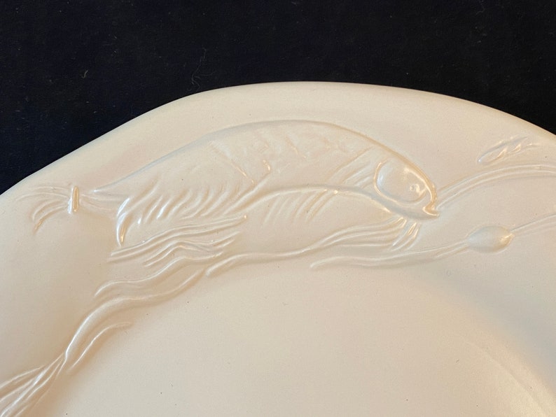 K&ouml;nnte beinhalten: Eine wei&szlig;e Keramik-Servierplatte mit einem erhabenen Design eines Fisches, der durch Wasser schwimmt. Der Fisch ist mit Schuppen und Flossen detailliert.