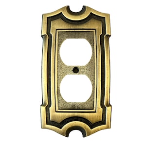 Puede incluir: Una tapa de toma de corriente dorada con un diseño decorativo y ornamentado. La tapa tiene una superficie texturizada en relieve y un borde negro.