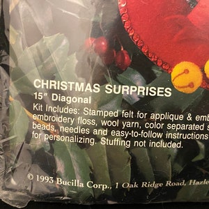 May include: A close-up of a Christmas craft kit with a red felt stocking and jingle bells. The kit includes stamped felt, embroidery floss, wool yarn, sequins, beads, needles, and instructions. The kit is 15 inches diagonally. &copy; 1993 Bucilla Corp., 1 Oak Ridge Road, Hazleton, PA 18