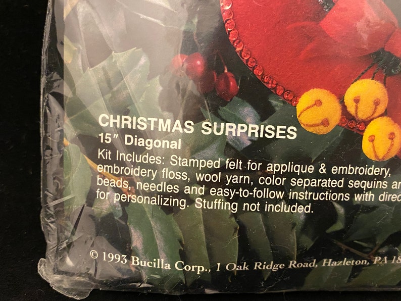 May include: A close-up of a Christmas craft kit with a red felt stocking and jingle bells. The kit includes stamped felt, embroidery floss, wool yarn, sequins, beads, needles, and instructions. The kit is 15 inches diagonally. &copy; 1993 Bucilla Corp., 1 Oak Ridge Road, Hazleton, PA 18