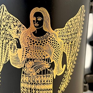 K&ouml;nnte beinhalten: Schwarze Tasse mit einem goldenen Design eines Engels mit Fl&uuml;geln. Der Engel tr&auml;gt ein gemustertes Kleid und h&auml;lt einen Blumenstrau&szlig;.