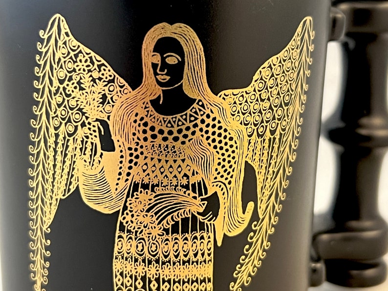 K&ouml;nnte beinhalten: Schwarze Tasse mit einem goldenen Design eines Engels mit Fl&uuml;geln. Der Engel tr&auml;gt ein gemustertes Kleid und h&auml;lt einen Blumenstrau&szlig;.