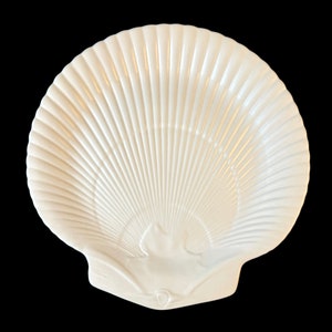 May include: A white ceramic dish shaped like a large scallop shell. The surface features a series of raised, radiating ridges that extend from the base to the scalloped outer edge, creating a textured, fan-like appearance with a glossy finish.