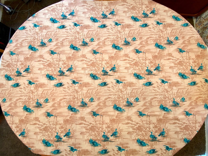 May include: A beige oval tablecloth with a repeating pattern of blue ducks swimming in a pond with brown reeds.