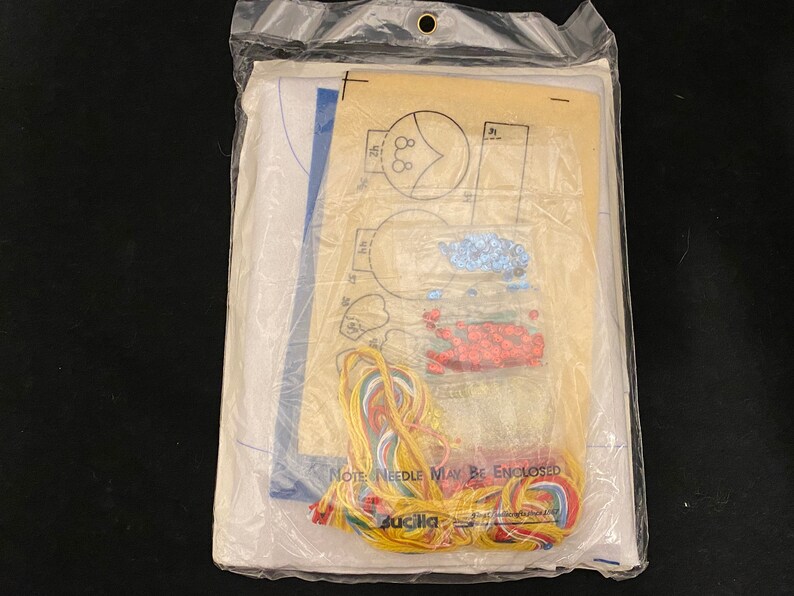 May include: A craft kit with a white fabric base, colorful embroidery floss, and a pattern for creating a beaded design. The kit includes instructions and a needle. The packaging includes the text "Note: Needle May Be Enclosed".