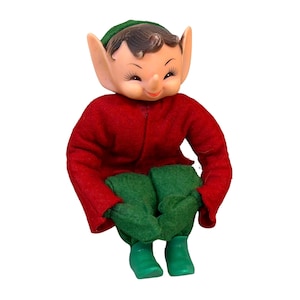Op de afbeelding: Een vintage elfpop met een gegoten kunststof gezicht, puntige oren, donker haar en een glimlachende uitdrukking. De pop draagt een rood vilten shirt met lange mouwen, een groene vilten broek, een groene vilten muts en groene kunststof schoenen.