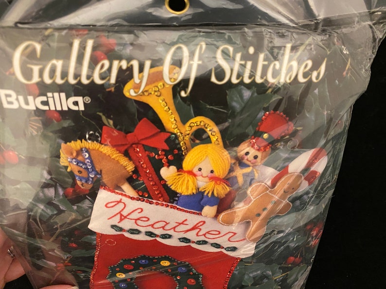 May include: A Bucilla Gallery of Stitches stocking kit featuring a red stocking with a green holly wreath, a gingerbread man, a toy soldier, a doll, a rocking horse, and a trumpet. The stocking is personalized with the name "Heather".